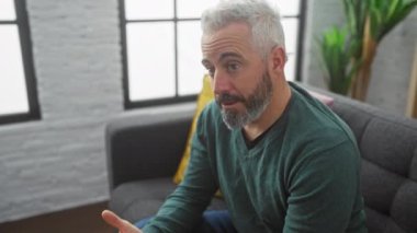 Oturma odasında sakallı bir adam. Çatı katı pencereli. Sohbet ve tefekkür ediyor..