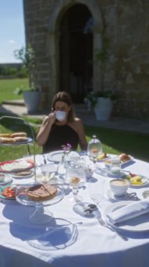 Genç bir kadın Puglia, İtalya 'da bir villada kahvaltıdan zevk alıyor. İçinde çeşitli hamur işleri, kruvasanlar, meyveler ve huzurlu bir bahçede kahve bulunan bir masa var..
