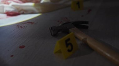 Eldivenli bir el, kasvetli bir suç mahallinde kanıt olarak işaretlenmiş kanlı bir çekici araştırır..