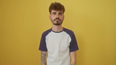 Genç İspanyol adam utanmış bir şekilde dikiliyor, örtülü elinden bakıyor, izole edilmiş sarı bir arka planda gözleriyle şoku ifade ediyor.