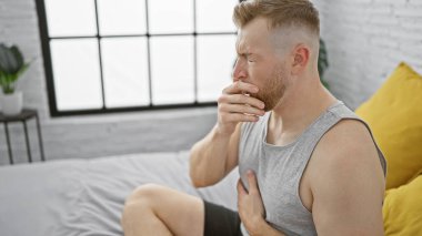 Sakallı, yorgun, genç bir adam yatak odasında oturup ağzını kapatıyor..