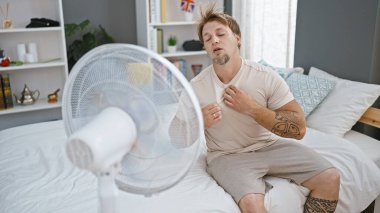 Pijamalı sakallı genç bir adam modern bir yatak odası ortamında bir hayranın önünde serinliyor..