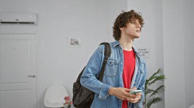 Kot ceketli genç bir adam sırt çantası ve akıllı telefonuyla parlak, modern bir ev ortamında dikiliyor, özenle yukarı bakıyor..