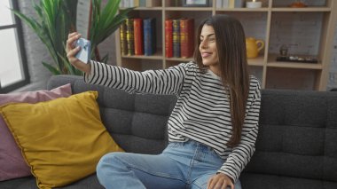 İspanyol kadın, oturma odasında akıllı telefonuyla selfie çekiyor.