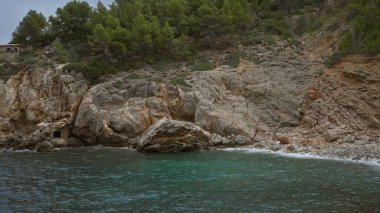 Bereketli yeşillik ile kayalık kıyı şeridi bulutlu bir gökyüzü altında turkuaz denizle buluşuyor dokunulmamış sahil doğasını ve engebeli araziyi açık havada gösteriyor.