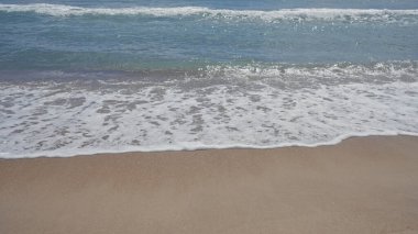 Kumsalda köpüklü dalgalar bulutsuz gökyüzünün altında yavaşça kıyıya vuruyor..