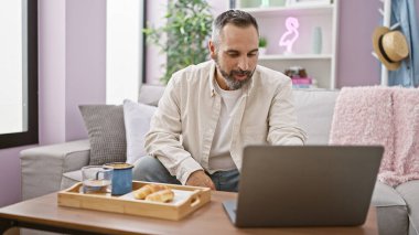 Yakışıklı, yaşlı, sakallı, gri saçlı bir adam rahat bir evde dizüstü bilgisayarla çalışıyor. Kanepe ve kahvaltı tepsisiyle..