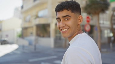 Şehir ortamında gülümseyen yakışıklı genç bir adam, açık havada neşe ve güven taşıyor..