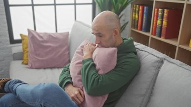 Sakallı, düşünceli, kel bir adam. Oturma odasındaki kanepede yastığı kucaklıyor..