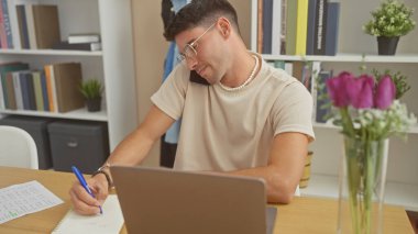 Yakışıklı, genç, İspanyol bir adam evde birden fazla iş yapıyor, notlar yazarken telefonla konuşuyor ve modern bir oturma odasında dizüstü bilgisayar kullanıyor..