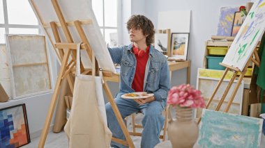 Bir sanat stüdyosunda resim yapan kıvırcık saçlı, kot ve kulaklık takan genç bir adam.