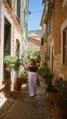 Fornalutx, Mallorca, İspanya 'da güneşli bir günde kırsal taştan binalar ve yemyeşil çimlerle çevrili pitoresk bir sokakta gezinen genç kadın.