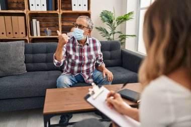 Sağlık maskesi takmış kıdemli bir adam yan tarafa parmağını uzatıp gülümsüyor ve psikoloji kliniğinde ağzı açık bir şekilde gülümsüyor.