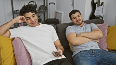 İki İspanyol adam, rahat bir odada, müzik aletleriyle birlikte kanepede dinleniyorlar..