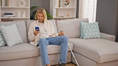 Sarışın adam akıllı telefon kullanıyor ve modern bir oturma odasında koltuk değneği tutuyor..