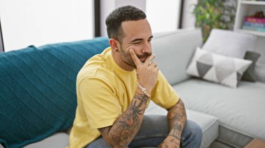 Modern bir oturma odasında oturan sakallı ve dövmeli dalgın bir adam rahat ve şık bir ortam yaratıyor..