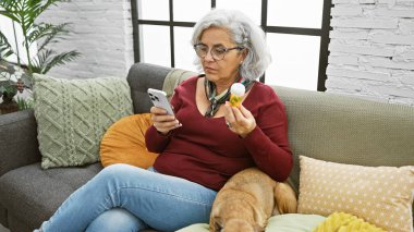 Akıllı telefon ve köpekle kanepede rahat bir oturma odasında oturmuş reçeteli ilaç şişesini inceleyen olgun bir kadın..
