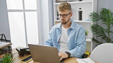 Modern kapalı ofis ortamında dizüstü bilgisayarla çalışan sakallı ve mavi gözlü yakışıklı beyaz adam.