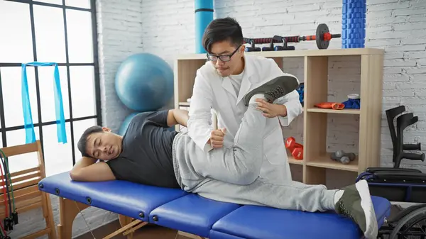 stock image A male physiotherapist assists a male patient with leg exercises in a well-equipped rehabilitation center.