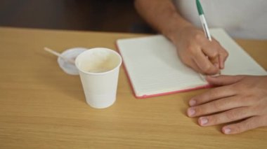 İspanyol bir adam, elinde bir fincan kahveyle ofis ortamında deftere yazı yazıyor. Ellerinin ve çalışma alanının detaylarına yakın plan çekilmiş..