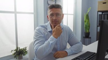 Ofis odasında oturan orta yaşlı bir adam, düşünceli bir ifadeyle poz veriyor, gözlük takıyor ve açık mavi bir gömlek giyiyor, masada bir bilgisayar ve arka planda bir bitki var..