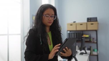 Modern bir ofiste tablet kullanan uzun siyah saçlı, gözlüklü, odaklanmış, masa ve pencerenin yanında duran bir kadın.