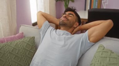 Evdeki kanepede uzanmış, elleri başının arkasında rahat bir oturma odasında dinlenen genç bir adam.