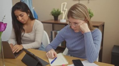 Evde çalışan ve akıllı telefon kullanan iki kadın, bir ev ortamında eğitim ve teknoloji sahnesini canlandırıyorlar..