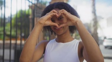 Kıvırcık saçlı, genç, Afro-Amerikan bir kadın. Elleriyle kalp şekli oluşturan ve gülümseyen bir kadın..