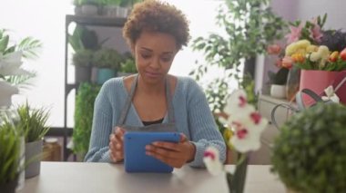 Çeşitli bitkiler ve renkli çiçeklerle çevrili bir çiçekçide tablet kullanan Afrikalı Amerikalı kadın..