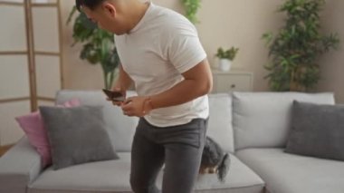 Oturma odasında genç bir adam akıllı telefon kullanarak küçük köpeğiyle etkileşim kuruyor. Bitkiler ve modern dekorlarla rahat ve rahat bir ortam sergiliyor..