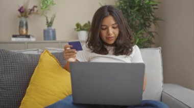 Genç bir kadın internetten kredi kartıyla alışveriş yapıyor, oturma odasındaki kanepede dinleniyor, etrafı bitkiler ve dekorlarla çevrili..