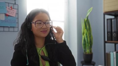 Ofis ortamında gülümseyen bir kadın, kulaklık ve gözlük takıyor, duvarında posterler, arka planda saksıda bir bitki, kendinden emin ve odaklanmış görünüyor..