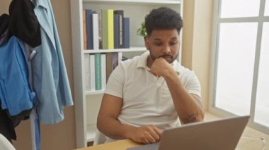 Ev ofisinde dizüstü bilgisayar, kitap rafı ve rahat kıyafetlerle çalışan Afrikalı Amerikalı bir adam..