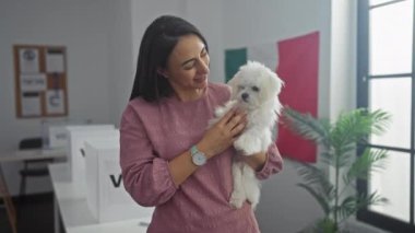 Meksika bayrağıyla modern bir ofiste beyaz bir bichon köpeğini tutan gülümseyen bir kadın..