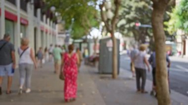 Güneşli bir şehir sokağında arka planda bulanık ağaçlarla yürüyen insanlar açık şehir ortamını gösteriyor.