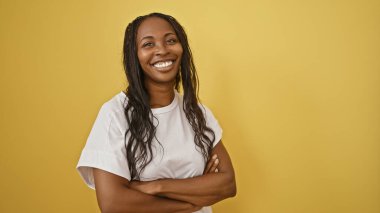 Confident woman with curly hair standing against a yellow background, arms crossed in a casual white shirt. clipart