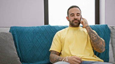 Sakallı ve dövmeli yakışıklı İspanyol bir adam evdeki turkuaz koltukta oturmuş, rahat bir şekilde kendine güveniyor..