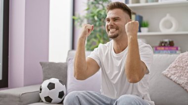 Neşeli, genç İspanyol bir adam modern bir salonda futbol topuyla otururken kutlama yapıyor..