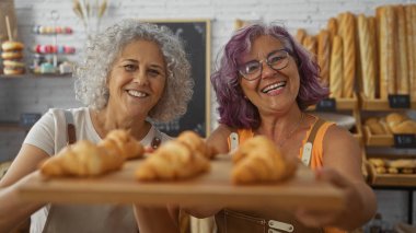 Gülümseyen orta yaşlı kadın fırıncılar arka planda çeşitli ekmek ve hamur işleriyle birlikte kapalı bir fırında taze kruvasan sunmaktan gurur duyarlar..