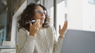 Orta yaşlı İspanyol bir kadın, modern ofis ortamında akıllı telefonuyla konuşuyor. Profesyonel iletişim duygusunu çağrıştırıyor..