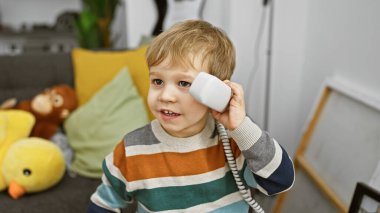 Küçük sarışın çocuk rahat bir oturma odasında telefonla oynuyor pelüş oyuncaklarla.