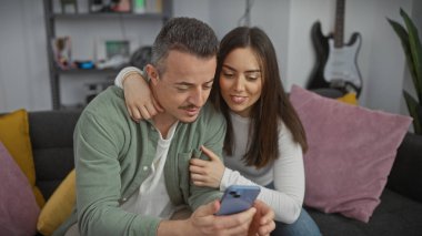 Sevgi dolu bir ilişki yaşayan kadın ve erkek, evlerinde birlikte vakit geçirmenin keyfini çıkarırlar. Oturma odasındaki akıllı telefona bakıp gülümserler..