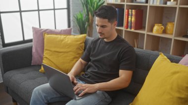 Odaklanmış, sakallı genç bir İspanyol, rahat bir oturma odasında kanepede otururken dizüstü bilgisayarla çalışıyor..