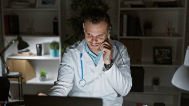 Beyaz önlüklü mutlu bir İspanyol erkek doktor gece klinikte telefonla konuşuyor..