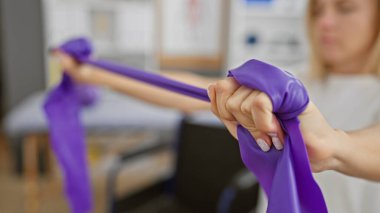 Odaklanmış genç bir kadın rehabilitasyon merkezinin bulanık iç kısmında mor bir direniş bandıyla egzersiz yapıyor..