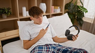 Kafkasyalı genç bir çocuk, elinde kulaklıkla yatak odasındaki telefonu inceliyor..