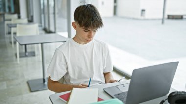 Modern bir kampüs kütüphanesinde dizüstü bilgisayar ve defterle çalışan odaklanmış genç bir adam..