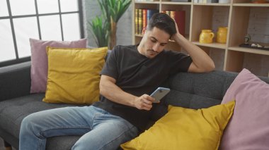 Rahat bir oturma odasında kanepede dinlenirken akıllı telefon kullanan düşünceli, sakallı genç bir adam..