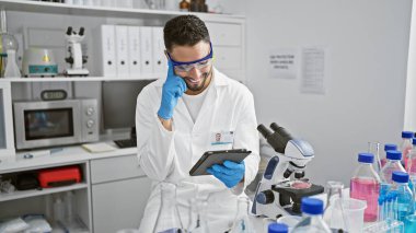 Laboratuvar önlüklü gülümseyen bir adam bilimsel ekipmanlarla dolu modern bir laboratuvarda dijital tablet kullanıyor..